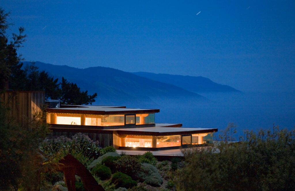 Sierra Mar at nightfall, its low glass-walled pavilions lit from within on the cliff edge while the Big Sur ridgeline dissolves into the sea behind.