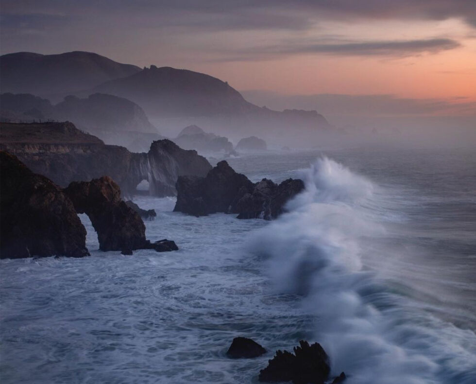 The Big Sur coastline at dusk, surf breaking against dark sea stacks and rock arches below fog-wrapped ridgelines that fade toward an apricot horizon.