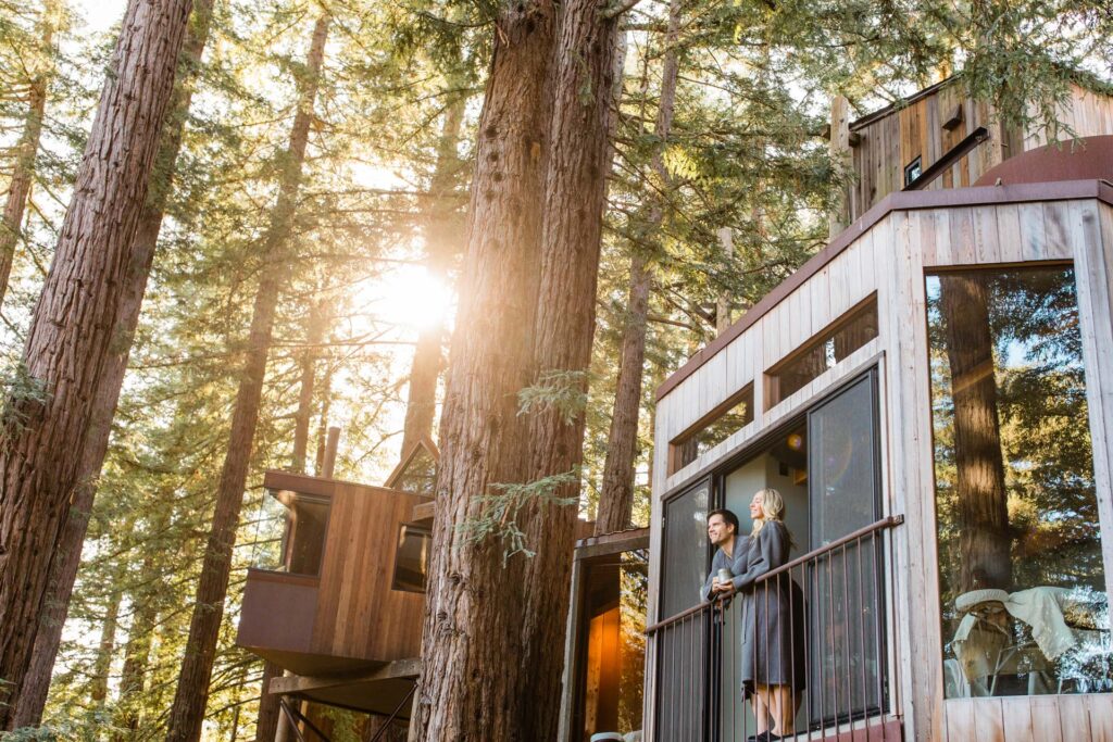 Two guests in robes taking morning coffee on the railed balcony of a Tree House, the sun flaring low through the redwood canopy around them.