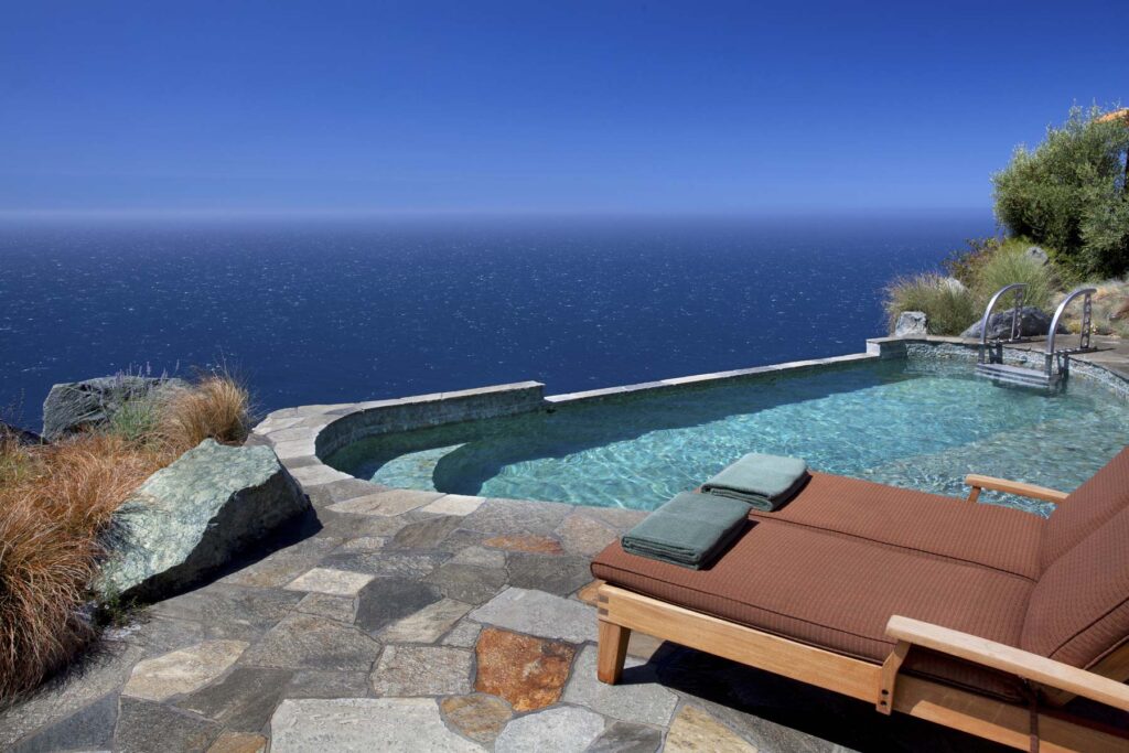 The Jade Pool on the cliff edge, shallow turquoise water behind a low stone rim, with a cushioned double lounger and folded green towels on the flagstone deck and open Pacific beyond.