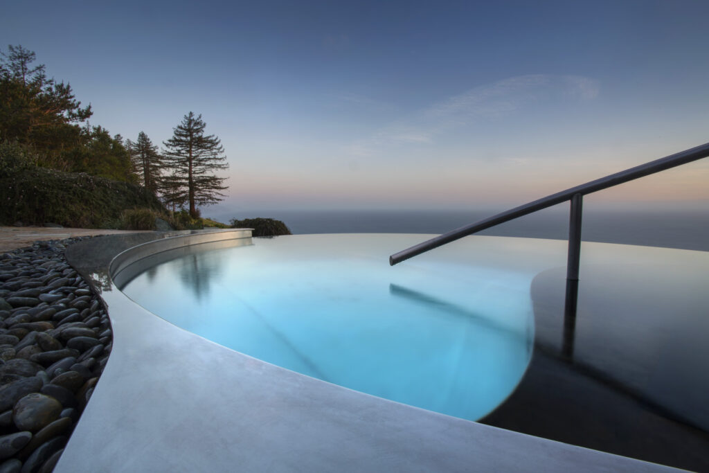 The infinity-edge Meditation Pool at dusk, pale blue water curving between a smooth concrete deck and a bed of river stones, its far edge running straight into the ocean sky.
