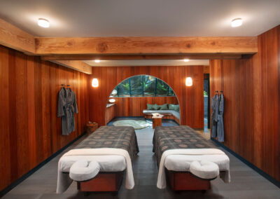 Spa treatment room with two massage tables, wooden paneled walls, robes on hooks, and warm lighting in a serene space.
