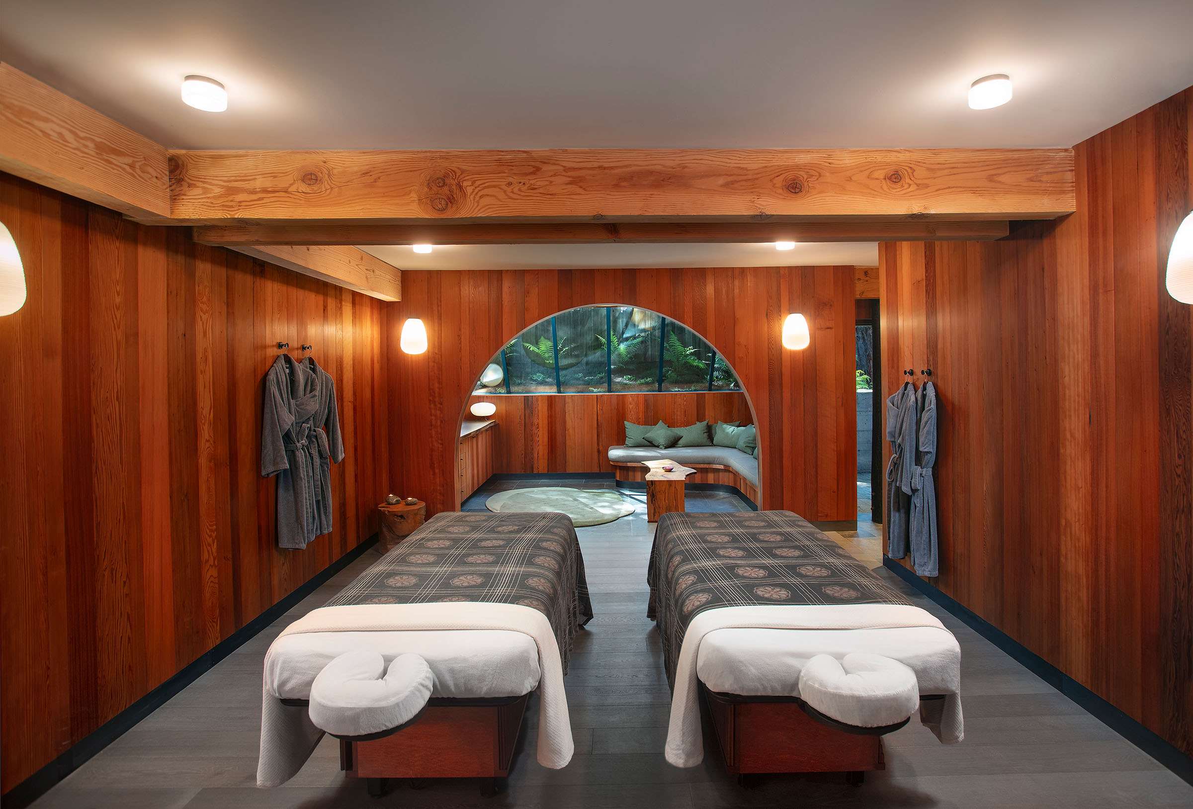Spa treatment room with two massage tables, wooden paneled walls, robes on hooks, and warm lighting in a serene space.