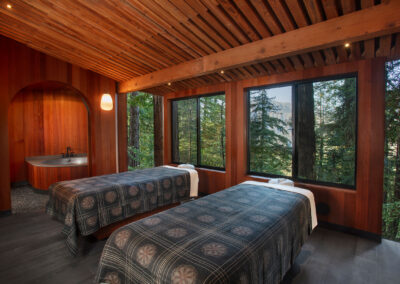 Wood-paneled couples spa treatment room with two massage tables, curved arch doorway with tub, and large forest-view windows revealing tall trees outside.