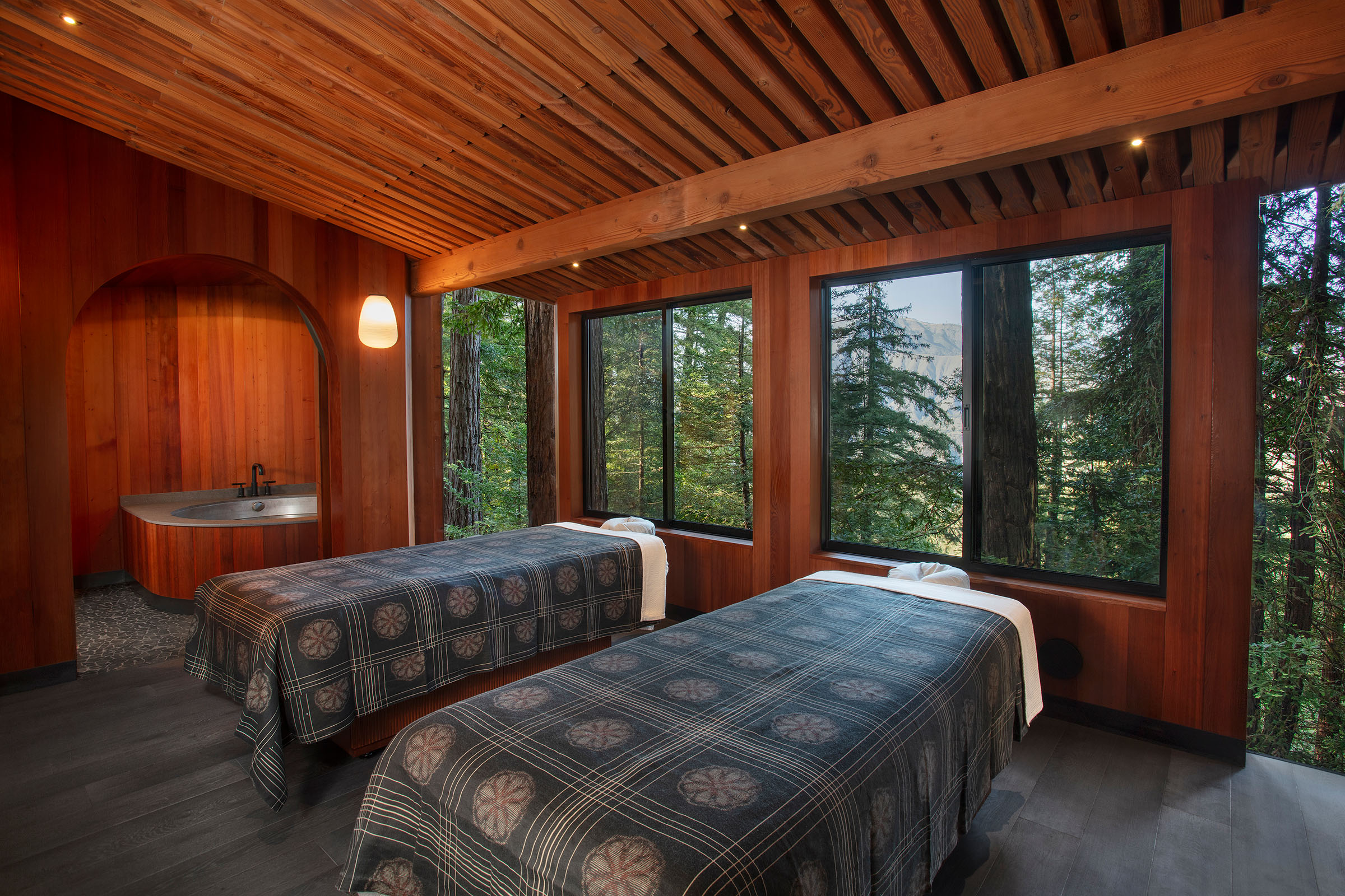 Wood-paneled couples spa treatment room with two massage tables, curved arch doorway with tub, and large forest-view windows revealing tall trees outside.