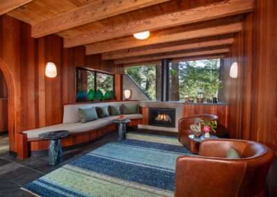 Wood-paneled living room with exposed ceiling beams, a fireplace, and windows overlooking trees outside.