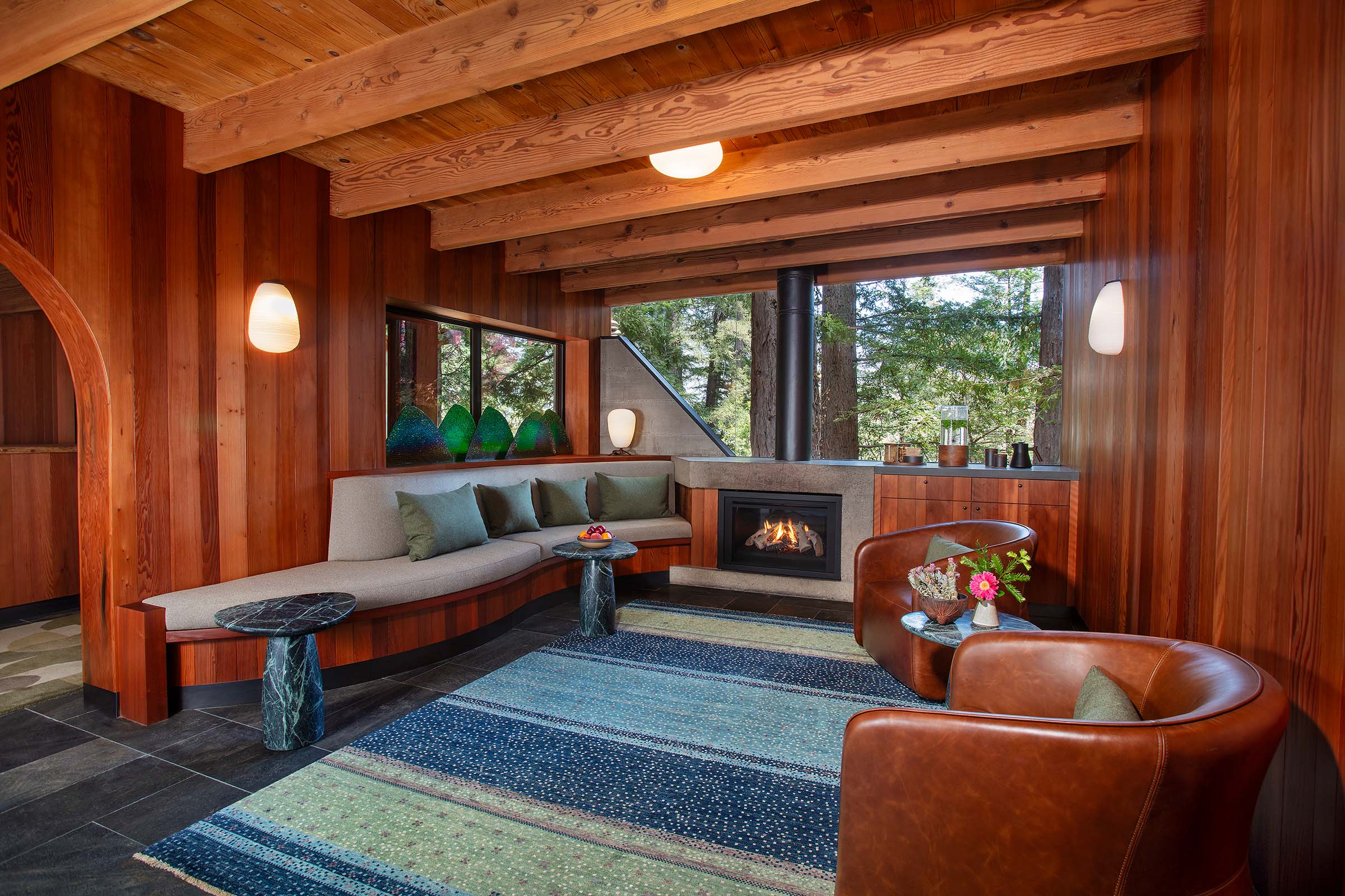 Wood-paneled living room with exposed ceiling beams, a fireplace, and windows overlooking trees outside.