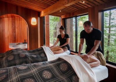 Two clients receive massages on adjacent beds in a wood-walled spa, with therapists working and forest visible through large windows.