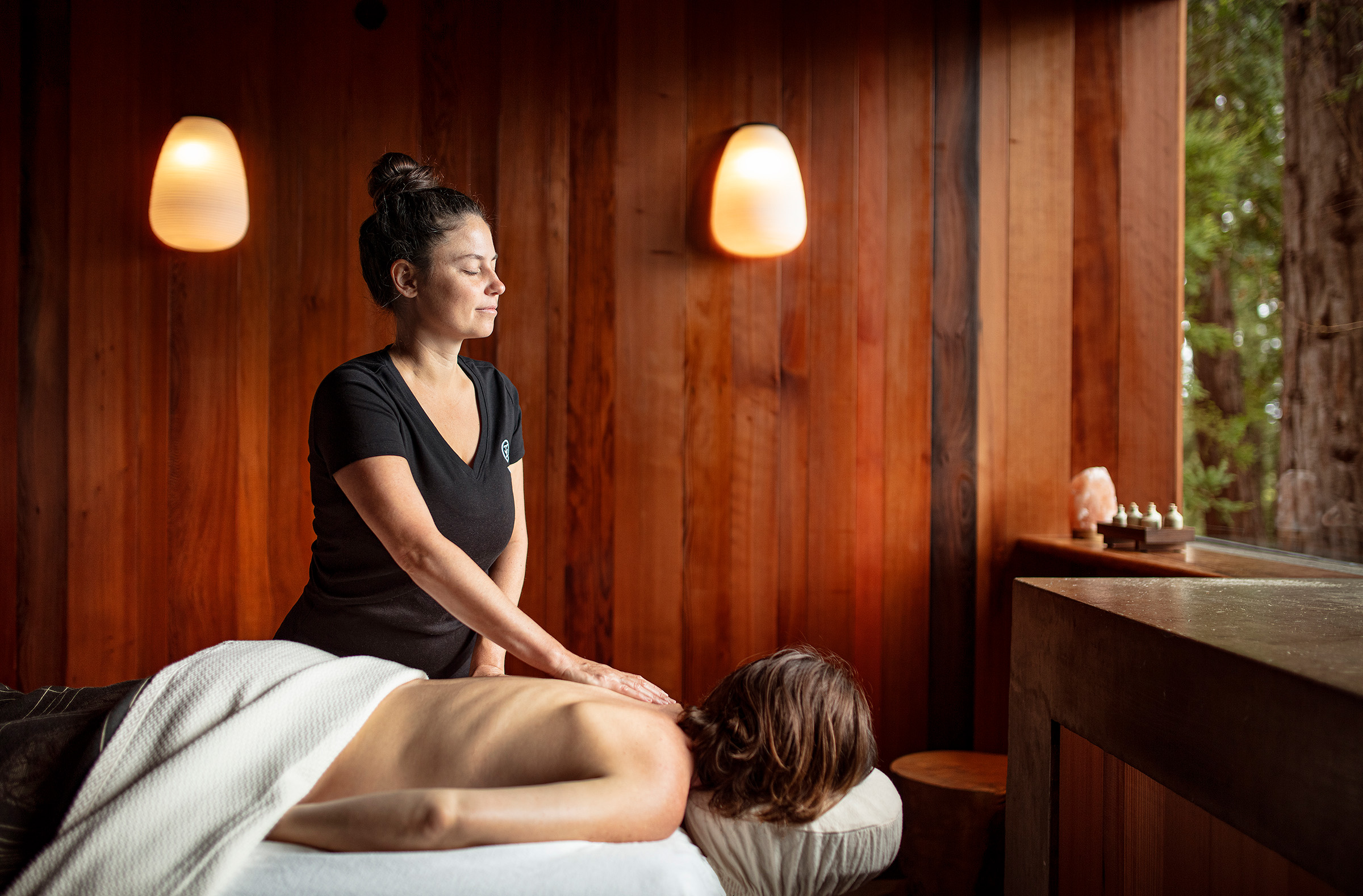 Spa massage in a warm, wood-paneled room: therapist standing beside a client lying face down on a massage table with a white towel.